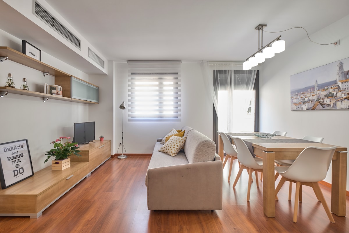 A modern living area features a comfortable sofa, a wooden entertainment unit with a TV, and a dining table accompanied by four chairs. Natural light filters through the windows, highlighting wooden flooring and minimalist decor, with a wall-mounted artwork depicting an urban scene.