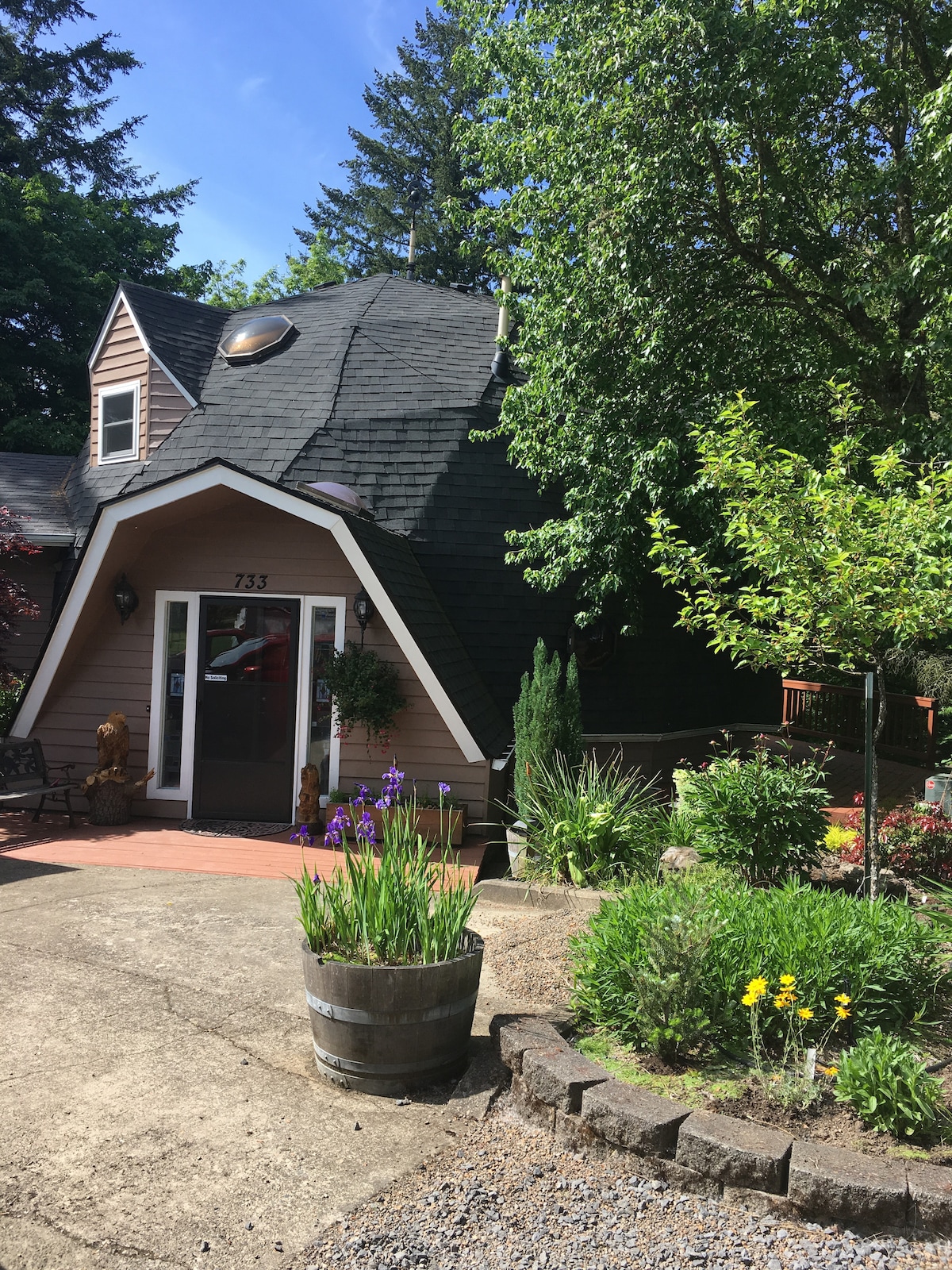 The exterior of a charming A-frame house is framed by lush greenery and colorful flowers. A welcoming entrance is visible, featuring a decorative door and bright landscaping in a tranquil outdoor setting.