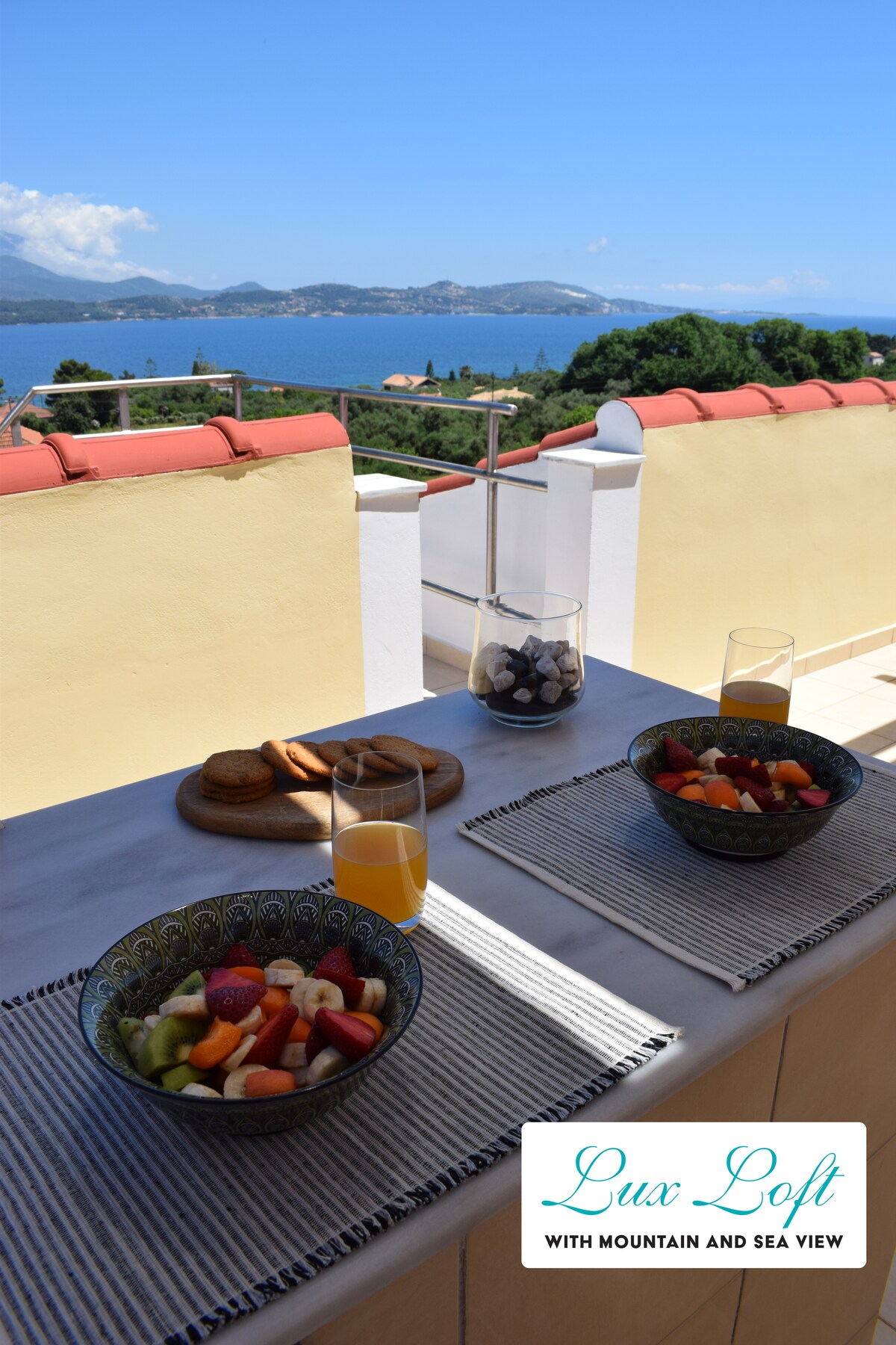 A balcony table is set with two bowls of fresh fruit and a glass of orange juice, complemented by additional snacks. The serene view of the sea and mountains is visible in the background, enhancing the tranquil setting.
