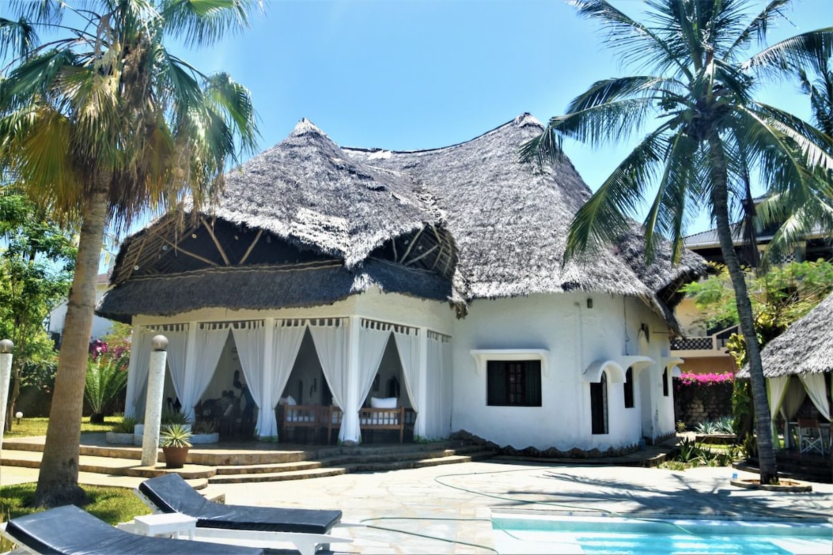 The villa features a thatched roof and an open terrace framed by palm trees. Soft white drapes sway gently in the breeze, offering a shaded seating area. A spacious pool area is visible, complemented by stone pathways and lush greenery surrounding the property.