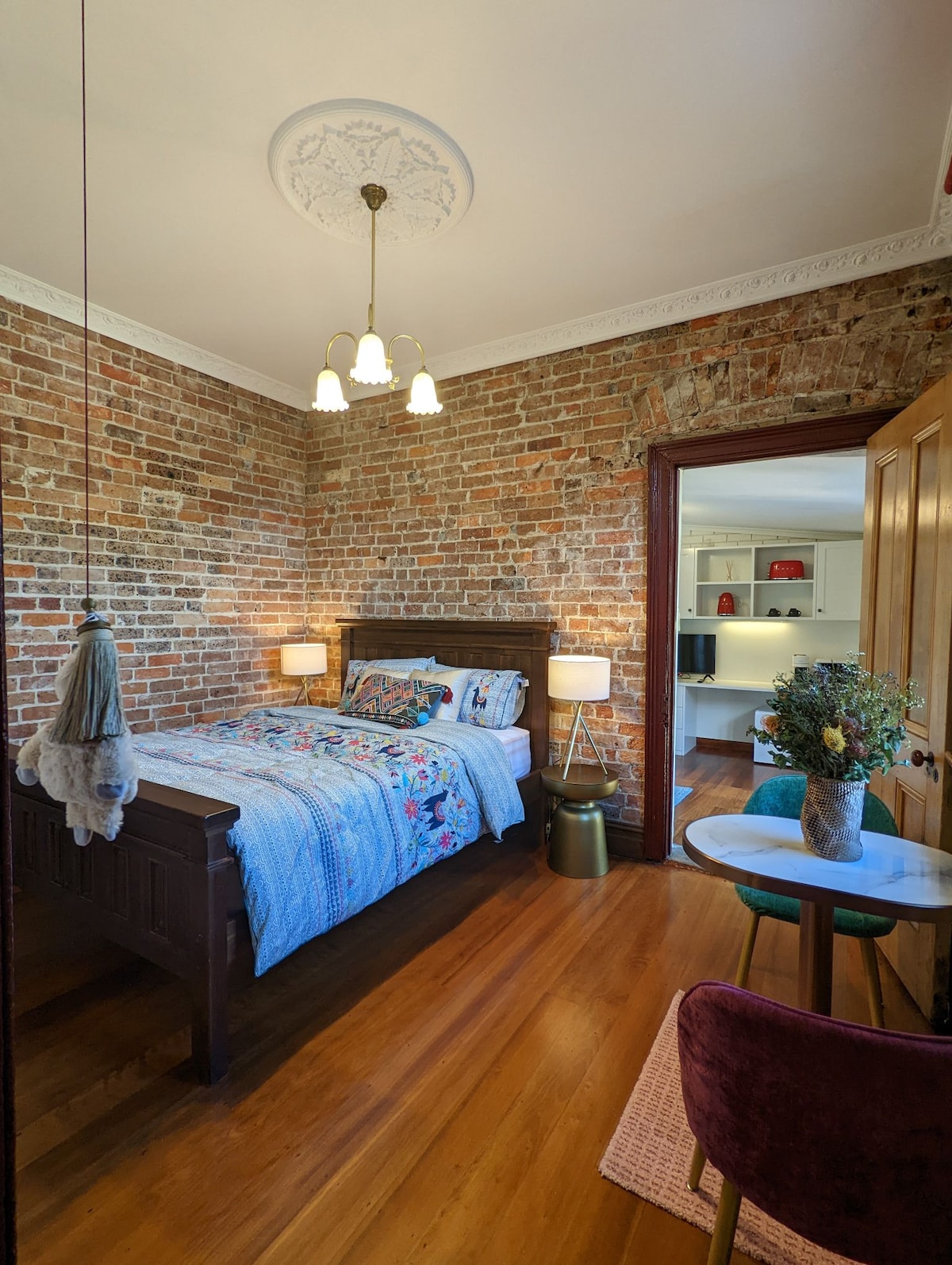 The guest room features an exposed brick wall, creating a warm backdrop for the wooden bed adorned with a colorful quilt. A small side table and chair complement the space, while soft lighting from pendant lamps creates a cozy ambiance. A desk area is visible in the background.