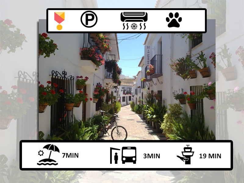 A charming, narrow street is lined with whitewashed buildings adorned with vibrant flower pots. A bicycle rests against a wall, while lush greenery complements the scene. The atmosphere is tranquil, reflecting the peaceful ambiance of the area, with convenient access to nearby amenities and attractions.