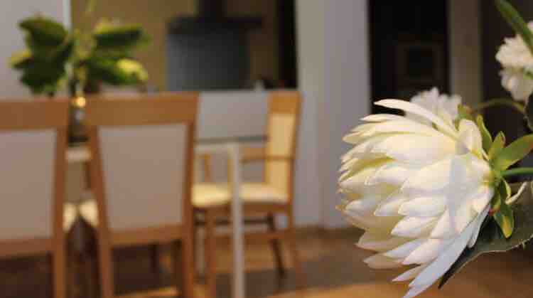 A dining area is visible with wooden chairs surrounding a table. A delicate white flower arrangement is prominently featured in the foreground, adding a touch of natural beauty. The background indicates a modern kitchen space with minimalist design.