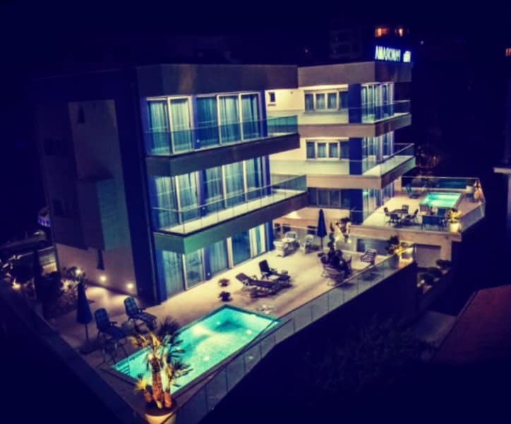 The luxurious villa is illuminated at night, revealing its three-story structure and multiple balconies. Two pools are visible on the terrace, surrounded by loungers and umbrellas. Lush vegetation complements the modern architecture, creating a serene outdoor space.