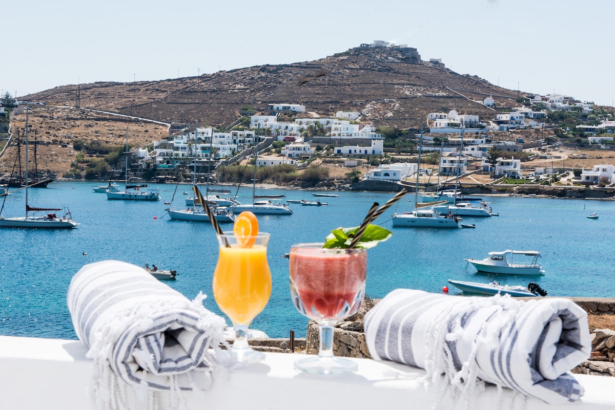 Two colorful cocktails are displayed on a table, surrounded by neatly rolled towels. In the background, a serene turquoise sea glistens with boats, while a scenic hillside dotted with white buildings creates a picturesque landscape.