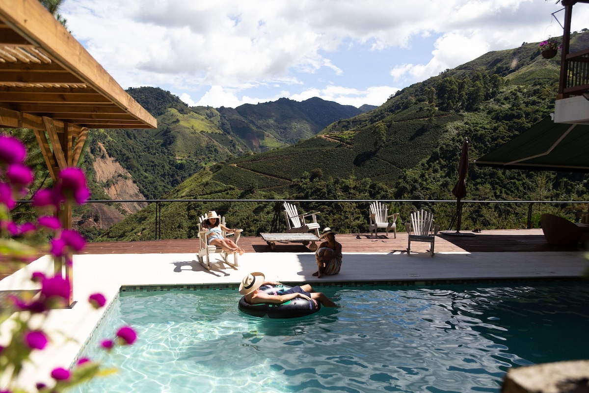 An outdoor heated pool is framed by a deck with lounge chairs, offering sweeping views of the surrounding mountains and lush coffee plantations. A person floats leisurely on an inner tube, with vibrant flowers adding color to the scene.