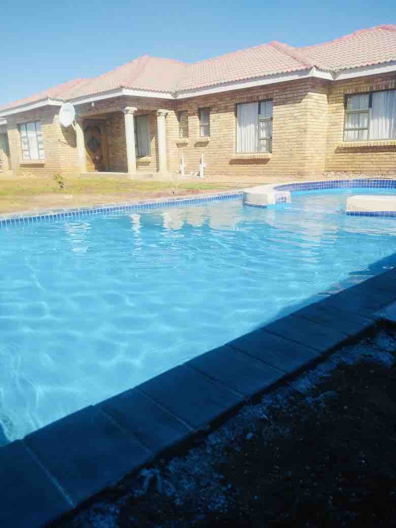 A clear blue pool reflects sunlight, creating a serene atmosphere. The surrounding area features a well-maintained lawn. A brick structure with a red-tiled roof can be seen in the background, providing an inviting backdrop for outdoor relaxation.