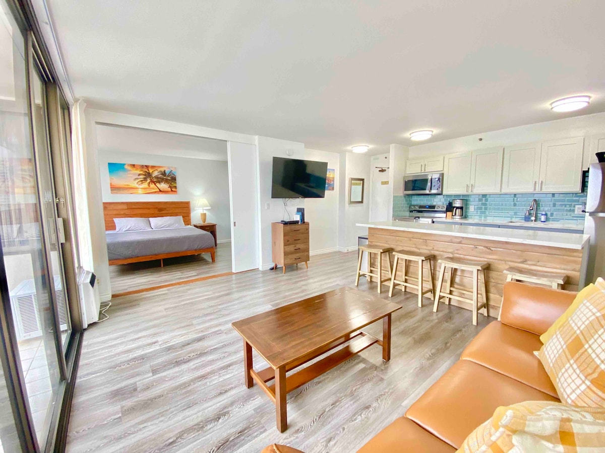 The open living space features a cozy seating area with light-colored furniture. A wooden dining island with stools is seen, along with a modern kitchen equipped with stainless steel appliances. A spacious bed is positioned in the background, accentuated by bright artwork and natural light.
