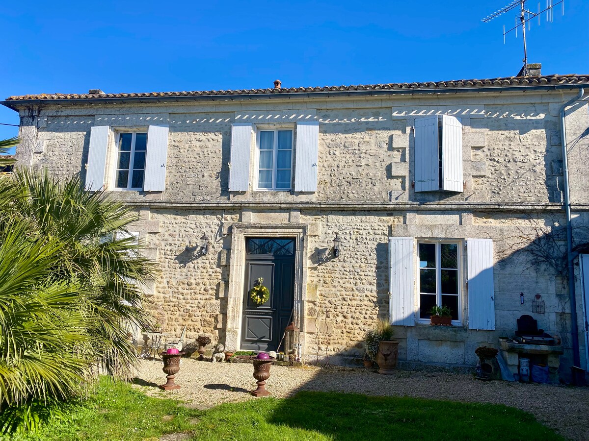The exterior of a charming stone house is presented, featuring traditional white shutters and a decorative front door. A landscaped area with green grass and potted plants is visible, alongside a gravel pathway leading to the entrance. Bright sunlight illuminates the facade.