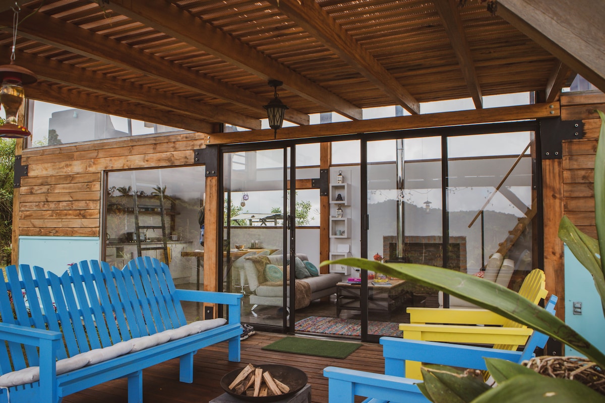 A covered terrace features bright blue and yellow seating, providing a cheerful outdoor lounge area. Large glass doors lead into the living space, where a cozy interior is visible. A fire pit is placed at the center, surrounded by greenery for a tranquil setting.