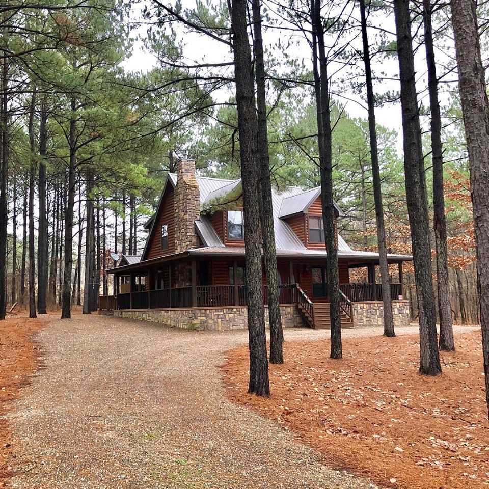 A spacious cabin is set among tall pine trees, showcasing a stone and wood exterior. The wrap-around deck, framed by the natural surroundings, invites relaxation. A gravel driveway leads to the entrance, surrounded by a carpet of pine needles and earthy tones.