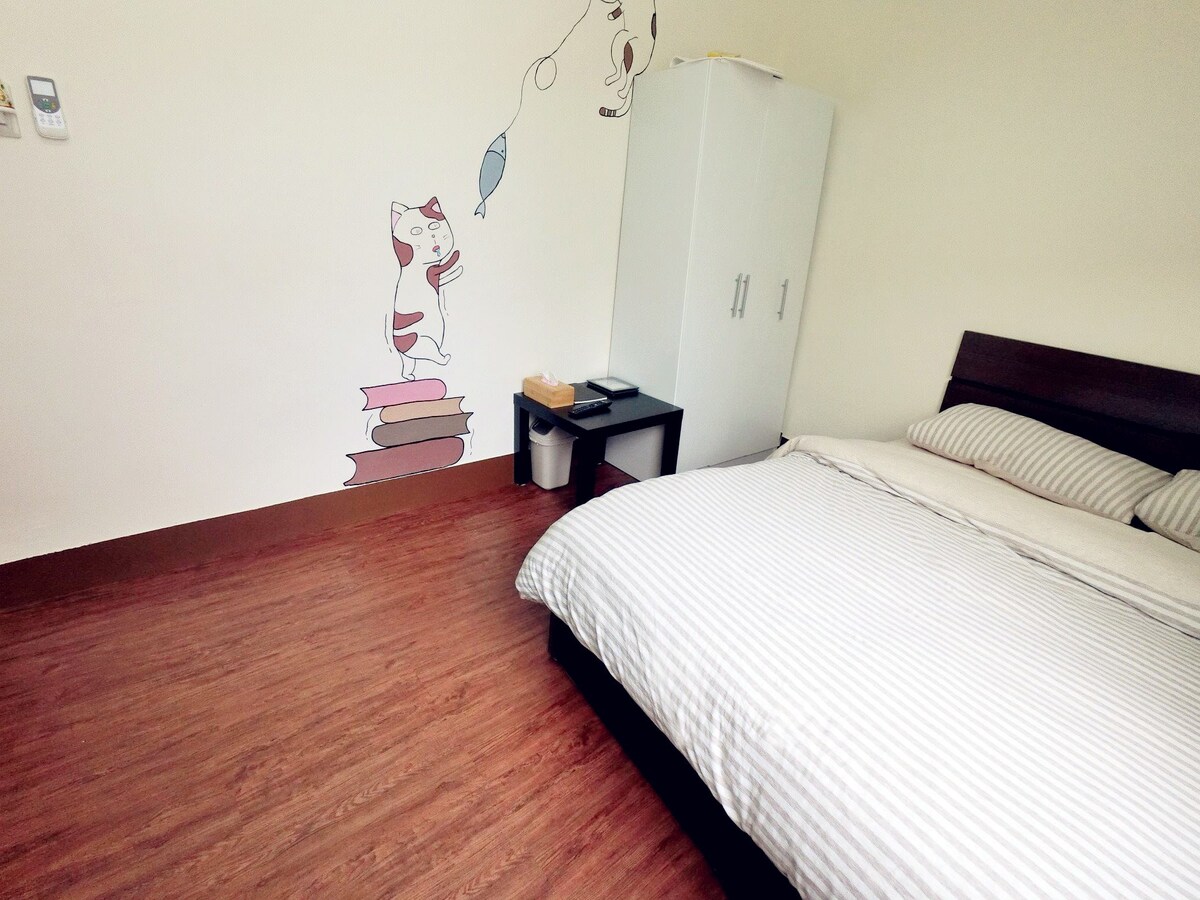 A quaint sleeping area features a spacious bed with striped bedding. A white cabinet is positioned in the corner, alongside a small table and wastebasket. A playful mural of a cat adds charm to the room's clean and simple design.