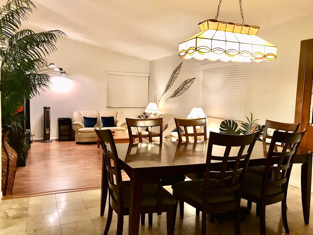 A spacious dining area is highlighted by a large wooden table surrounded by six chairs. Soft lighting from a pendant lamp creates a cozy ambiance. In the background, a comfortable living space with a sectional sofa, two lamps, and decorative plants is visible.