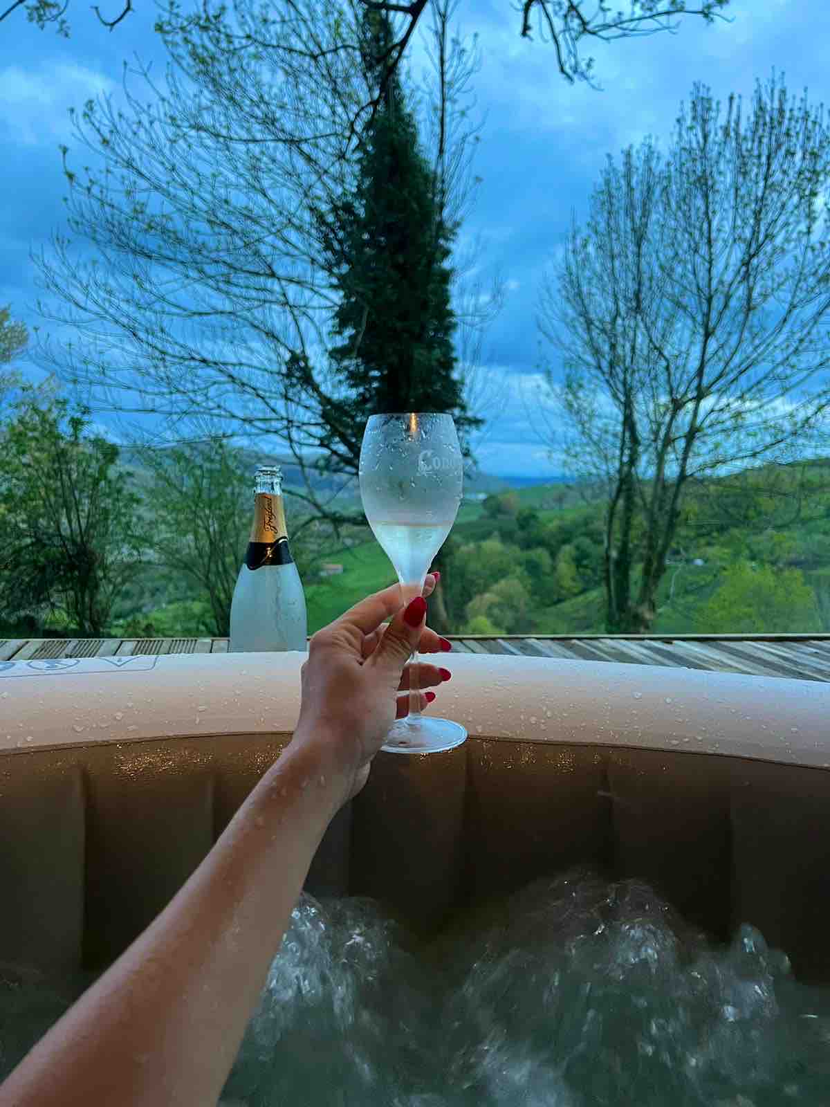 A bubbling jacuzzi is visible, set against a backdrop of lush greenery and a cloudy sky. A glass of sparkling drink is held above the water, while a bottle sits nearby, emphasizing a relaxing atmosphere surrounded by nature.