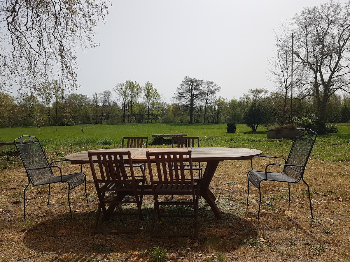 An outdoor dining area features a large wooden table surrounded by several wooden and metal chairs. The space overlooks a green lawn framed by trees, creating a serene environment for meals or gatherings.