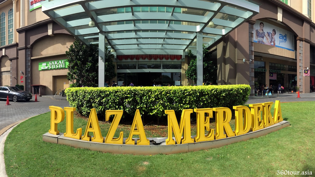 The entrance of Plaza Merdeka is prominently featured, with a large yellow signage displaying the name. A canopy stretches overhead, providing shade, while greenery and landscaped lawns enhance the welcoming atmosphere. The surrounding area includes shops and a smooth paved pathway for easy access.
