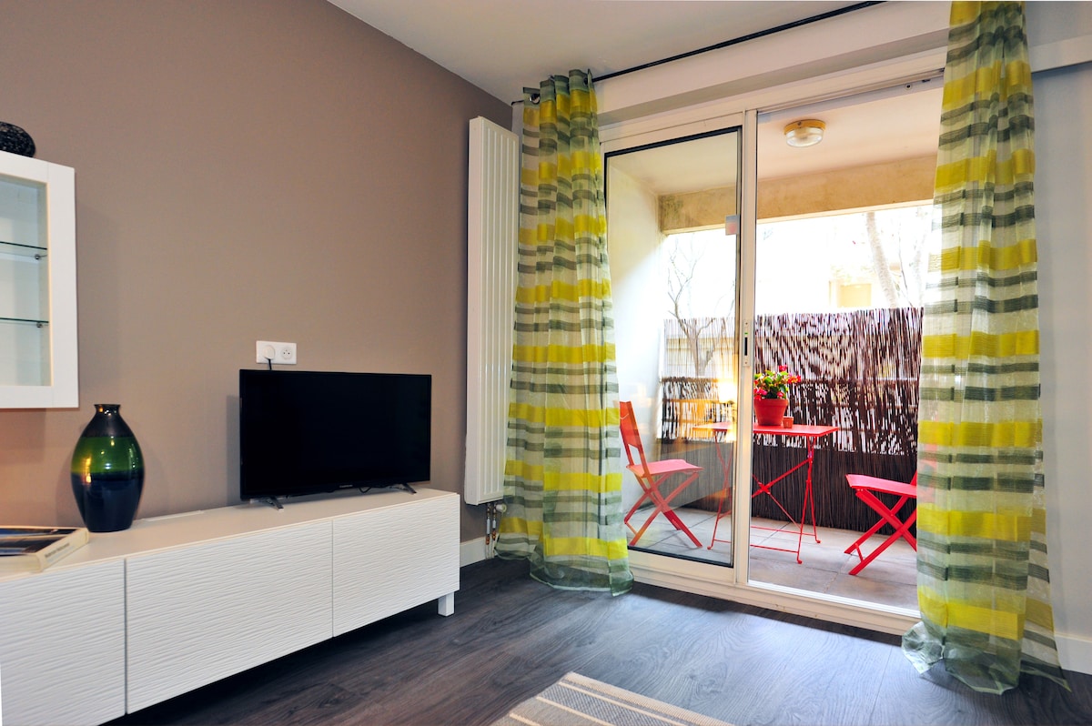 A modern living area features a sleek white TV unit with a television. Sheer curtains in a yellow and green pattern frame sliding glass doors, leading to a small balcony with two red chairs. Light wood flooring complements the neutral wall color.