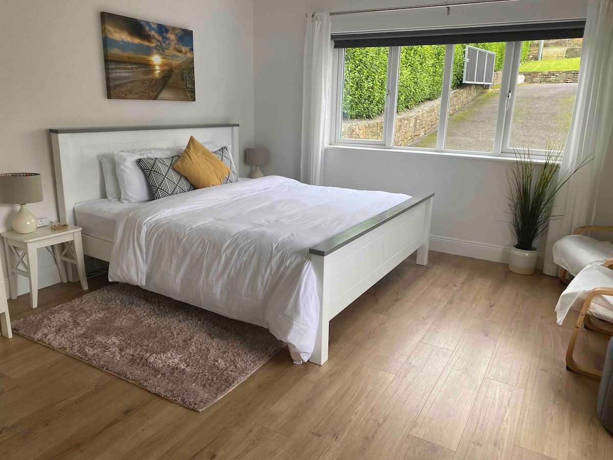 A spacious bedroom is furnished with a large bed topped with white linens and accented by a yellow pillow. Natural light enters through large windows, illuminating the wood flooring. A small side table and a chair are positioned in the room, adding to the serene ambiance.