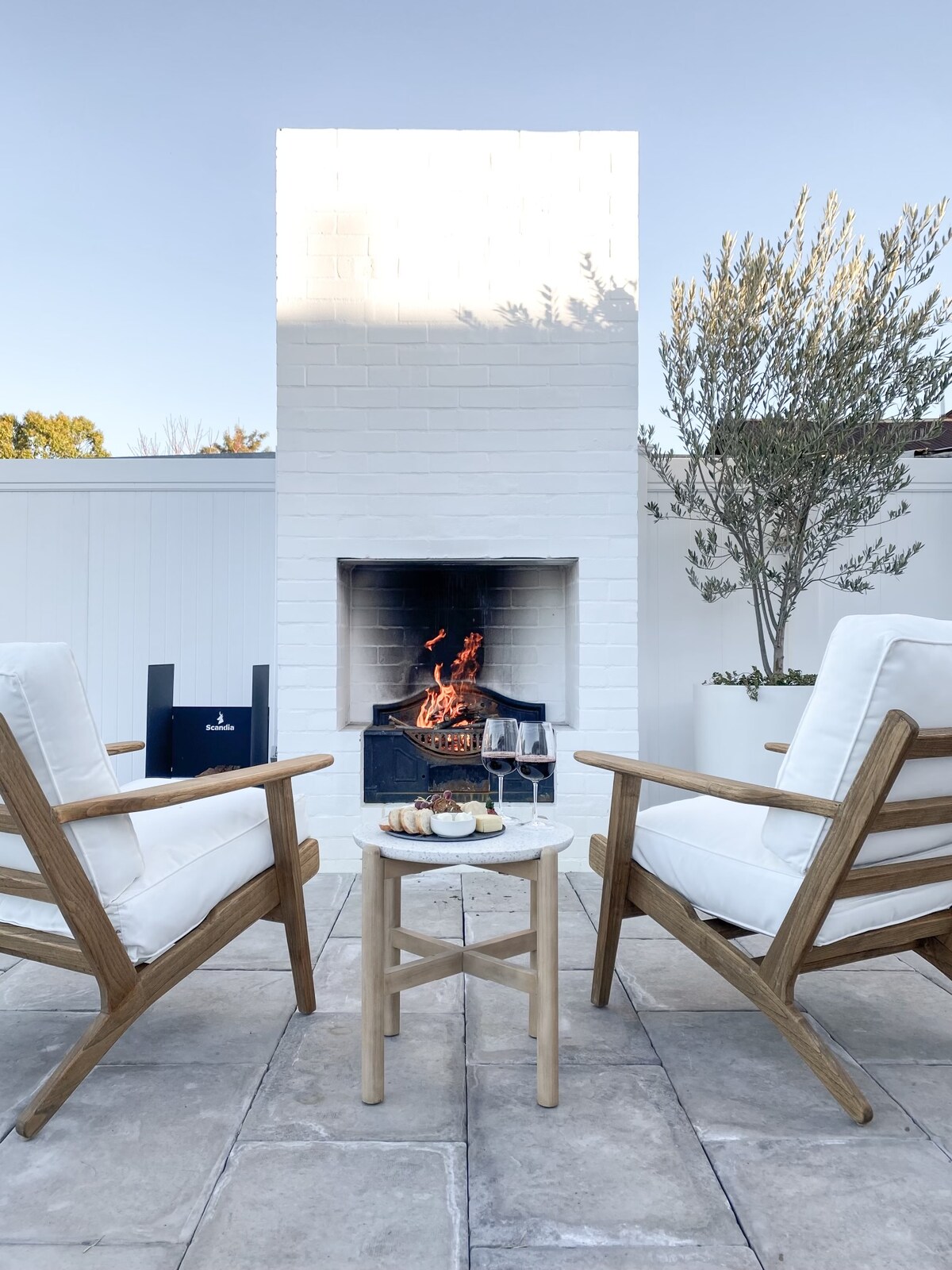 A cozy outdoor seating area is set before a white-brick fireplace, which features a gentle flame. Two cushioned chairs with wooden frames face a small table holding glasses of wine and a platter of snacks, surrounded by neatly arranged stone tiles and greenery.