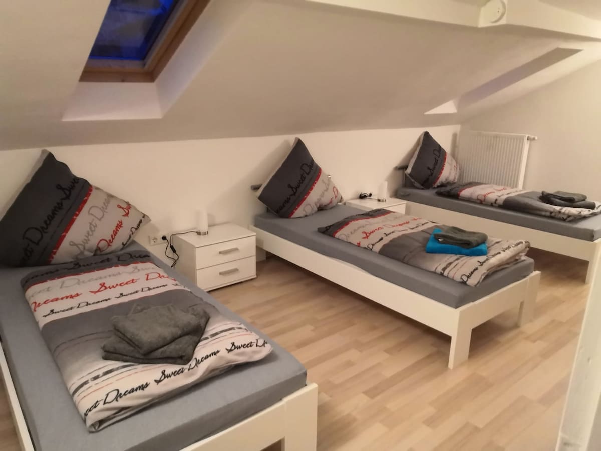 The room features three neatly arranged single beds, each with gray bedding and decorative pillows. A small bedside table is positioned next to one bed. Natural light enters through a skylight, illuminating the light wooden floors and the bright white walls.