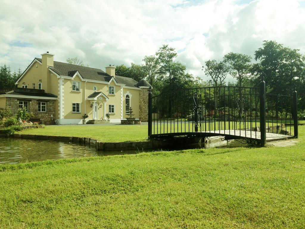 A charming B&B is set against a lush green lawn, featuring a stone bridge that spans a tranquil pond. The exterior showcases a blend of cream-colored stone and soft-colored walls, framed by trees and gentle landscaping under a partly cloudy sky.