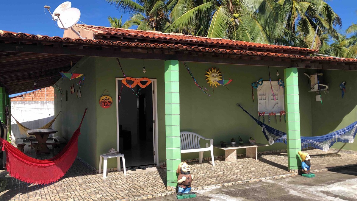 The exterior of a single-story house is shown, featuring a terracotta roof and green walls. Hammocks are hung on the porch, with tropical plants surrounding the area. A dining table is visible under the roof, and decorative elements, including figures and art, add character to the space.