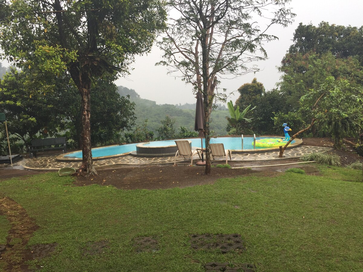 A serene outdoor area features a swimming pool surrounded by lush greenery. Comfortable lounge chairs are positioned near the water. The landscape showcases a gentle slope leading to distant hills, providing a peaceful backdrop with trees scattered throughout the setting.