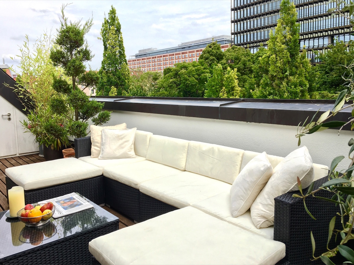 An inviting rooftop terrace features a large seating area with white cushions and black wicker furniture. A low table holds a bowl of fruit and a magazine. Lush greenery surrounds the space, while views of nearby buildings and trees provide a calming backdrop.