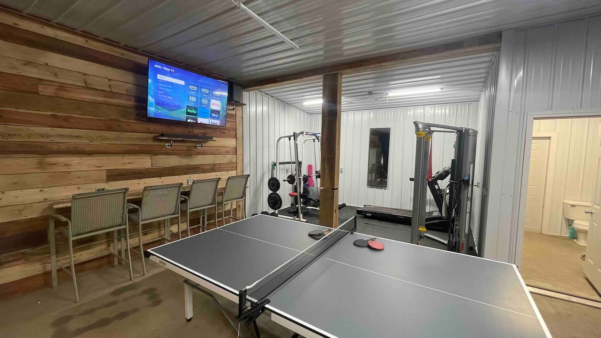 A multi-functional game room features a ping pong table in the foreground, with a TV mounted on the wall displaying options. Workout equipment and a small seating area are visible in the background, showcasing a modern layout with a metal and wood design.