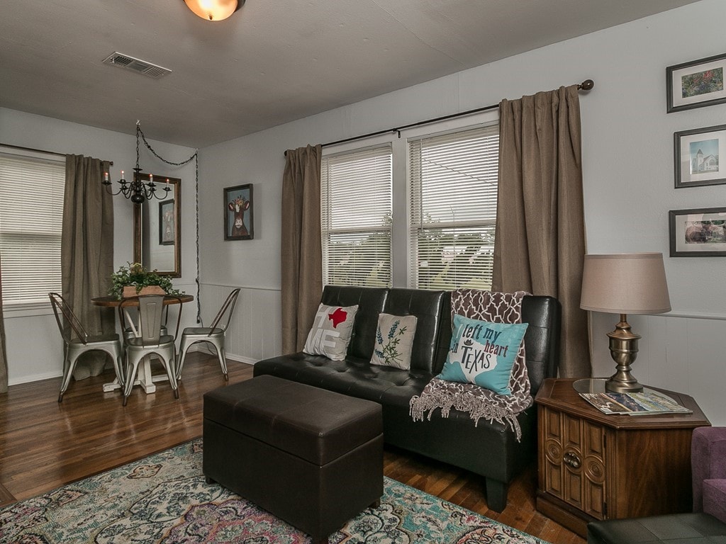 A cozy living area features a dark sofa with decorative pillows and a coffee table. Natural light streams through two windows, complemented by soft curtains. A dining area with a round table and metallic chairs is visible, adding to the inviting space.