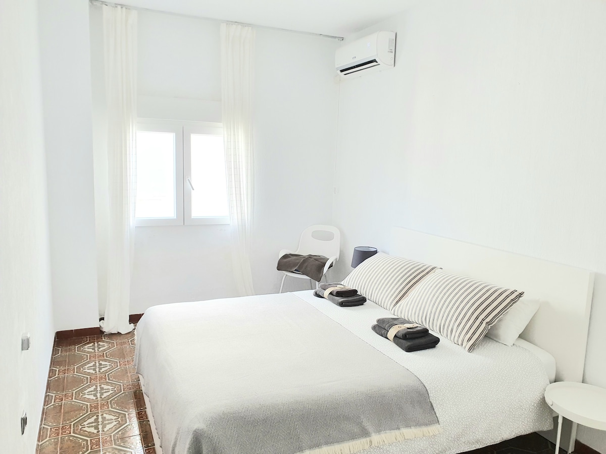 A bright room features a spacious bed with neutral bedding and decorative pillows. Natural light enters through a window adorned with sheer curtains. A small chair and a bedside table are visible, alongside a wall-mounted air conditioning unit.