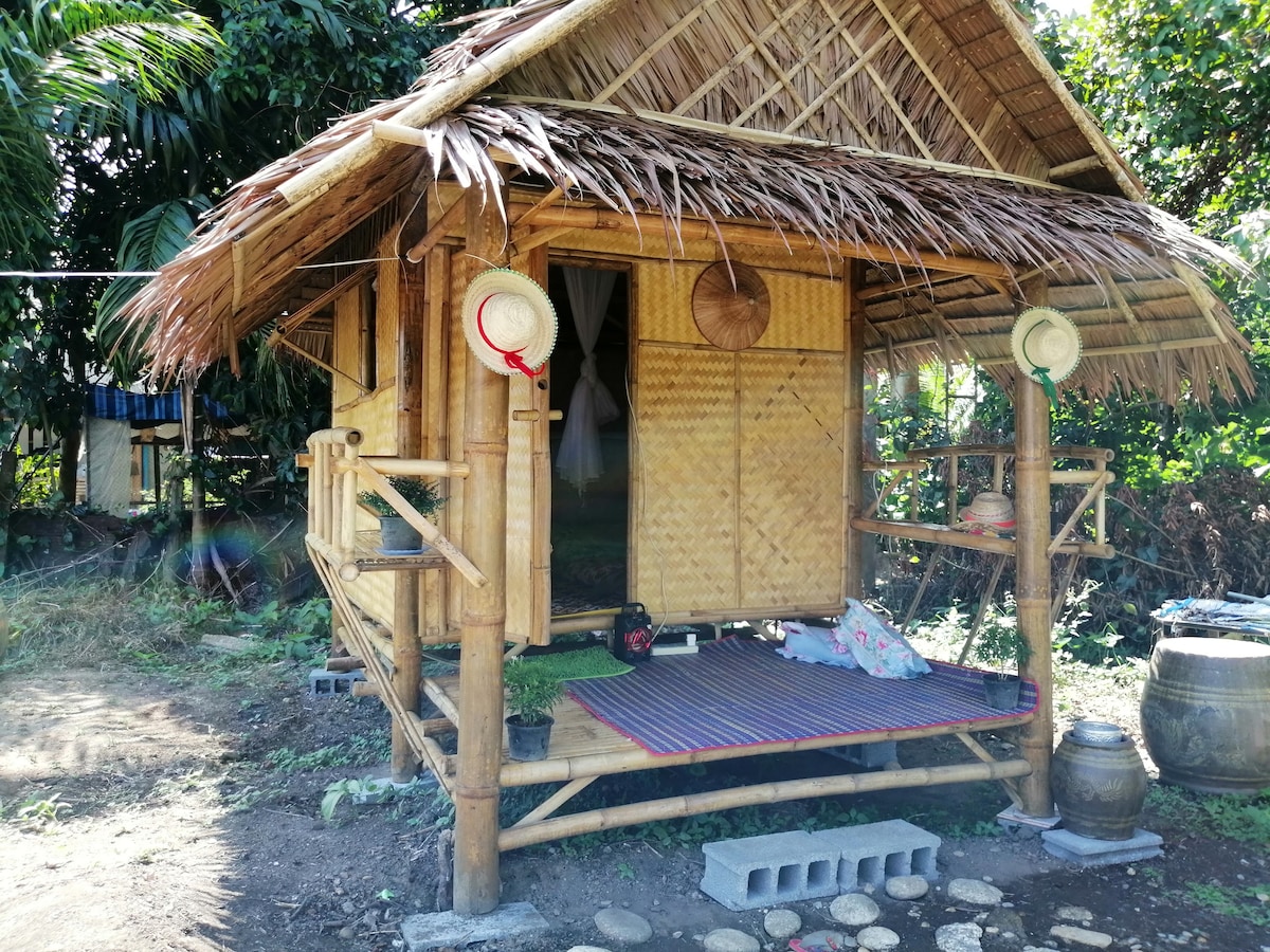 A bamboo hut surrounded by greenery features a thatched roof and a porch area. Two hats hang on bamboo supports, and a woven mat is laid on the floor. Natural materials create a simple yet inviting ambiance, showcasing the connection to the outdoors.