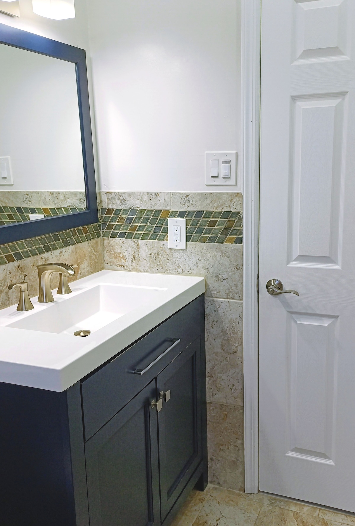 A modern bathroom features a sleek white sink atop a dark cabinetry with brushed silver fixtures. A mirrored cabinet is mounted above the sink, surrounded by light-colored tiles. A light green mosaic backsplash adds a subtle pop of color, while a white door leads to another space.
