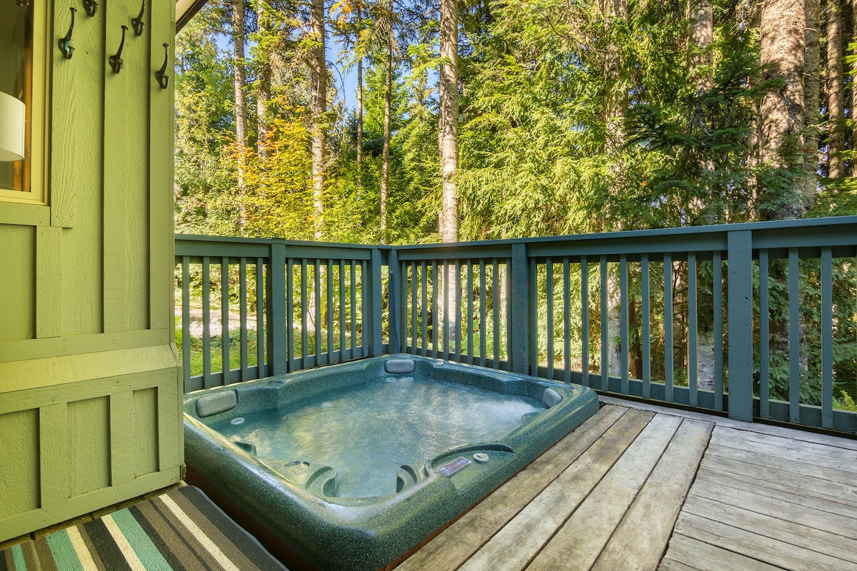 A private hot tub is positioned on a wooden deck, surrounded by tall trees. Sunlight filters through the foliage, creating a peaceful atmosphere. The tub features built-in seats and controls, providing a relaxing outdoor retreat.