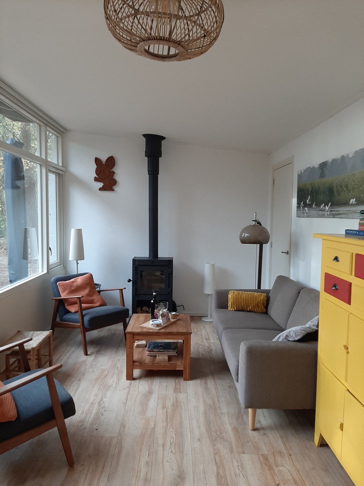 A living area is designed with large windows allowing natural light to enter. A wood-burning stove is positioned near a comfortable sofa and chairs, adorned with colorful cushions. A wooden coffee table sits in the center, complemented by a yellow storage unit against the wall.