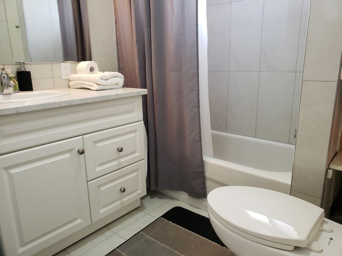 The bathroom features a clean white vanity with storage drawers and a marble countertop. A bathtub with a shower curtain is present alongside a modern toilet. Fresh towels are neatly arranged, and natural light filters in through the window, highlighting the tiled walls.