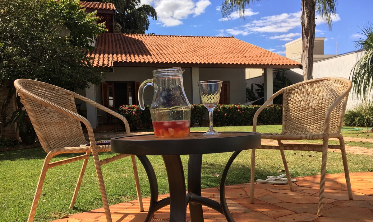 A small outdoor table is positioned on a stone patio, accompanied by two woven chairs. A glass pitcher filled with a beverage and a glass are placed on the table. Lush green grass surrounds the area, with a view of the house and palm trees in the background.
