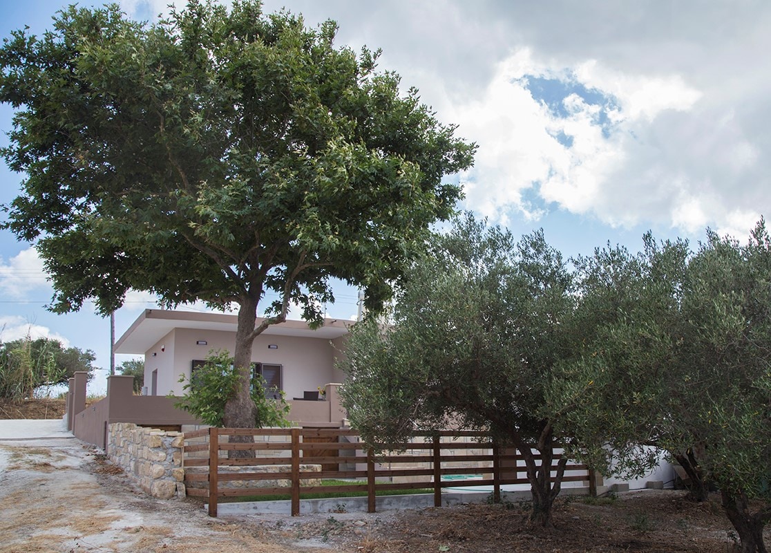 A modern house is framed by a large tree and olive trees in the foreground. The structure features a light-colored facade and is set against a cloudy sky. A wooden fence outlines the property, providing an inviting entrance.