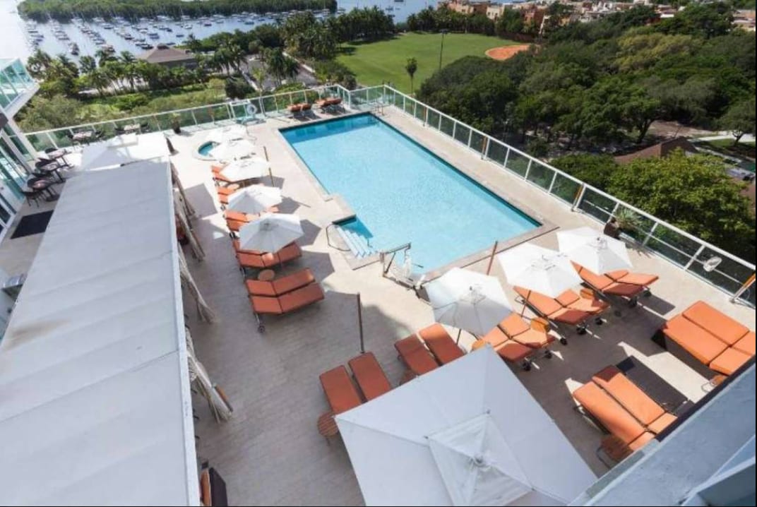 An aerial view of a rooftop pool area showcases a large swimming pool surrounded by lounge chairs with orange cushions. Several umbrellas provide shade, and greenery is visible in the background, alongside a marina and recreational fields.