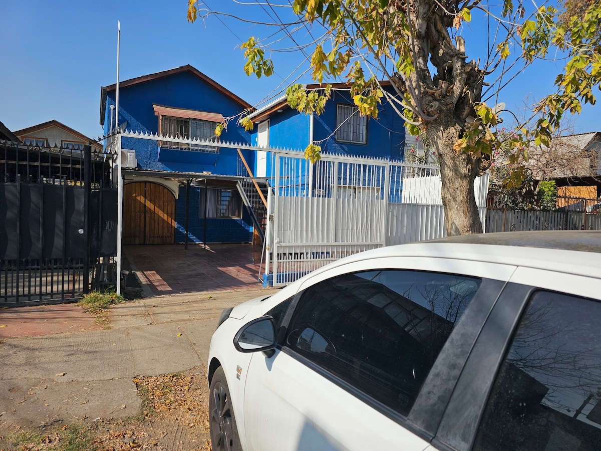 A blue house with a gated entrance is visible, showcasing a spacious front area bordered by a white fence. A tree with green leaves stands in the foreground, while a white vehicle is parked along the side of the road.