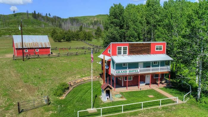 The Grand Hotel replica showcases a colorful exterior with red and blue accents. A spacious lawn surrounds the building, complemented by nearby trees and rolling hills. A red barn is visible in the background, adding to the rural charm of the property.