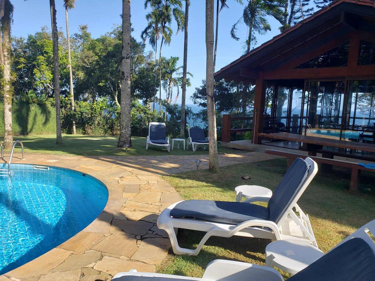 A private outdoor area features a sparkling pool surrounded by lush greenery. Sun loungers are arranged nearby, offering comfortable seating options. A covered terrace is visible in the background, providing shaded space with wooden seating and a view of the scenic landscape.