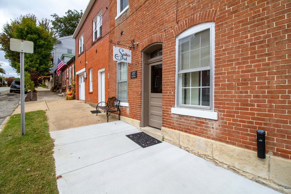 The exterior of a brick building is depicted, showcasing a welcoming entrance with a brown door. A small, black bench is positioned beside the door, and a concrete pathway leads to the entrance. Nearby, a flag can be seen adorning the neighboring building.