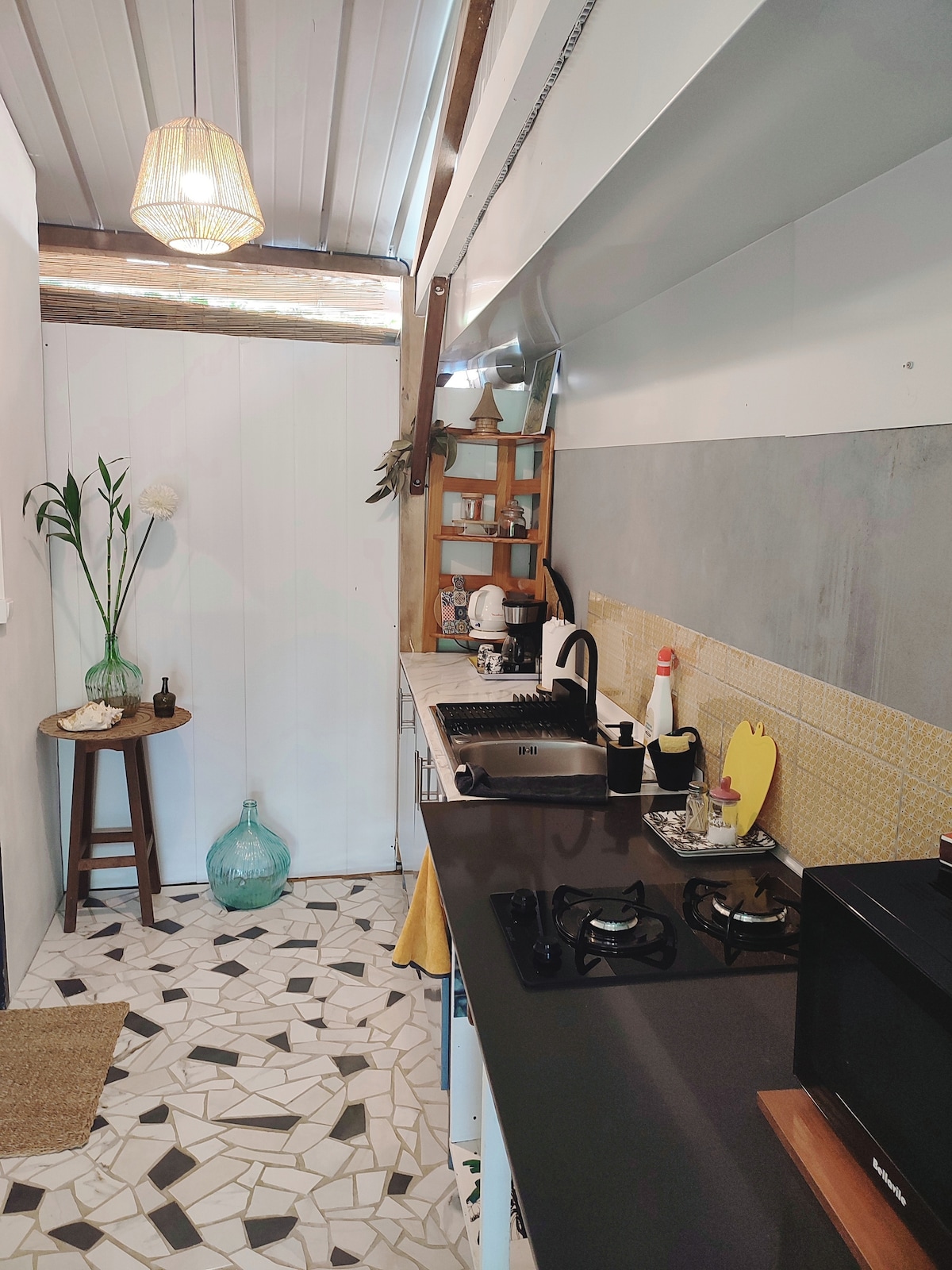The kitchen area features modern appliances, including a coffee maker and a stovetop. A decorative piece with a plant sits on a small wooden stool, while a large blue glass bottle adds a pop of color. The floor showcases an intricate mosaic design.