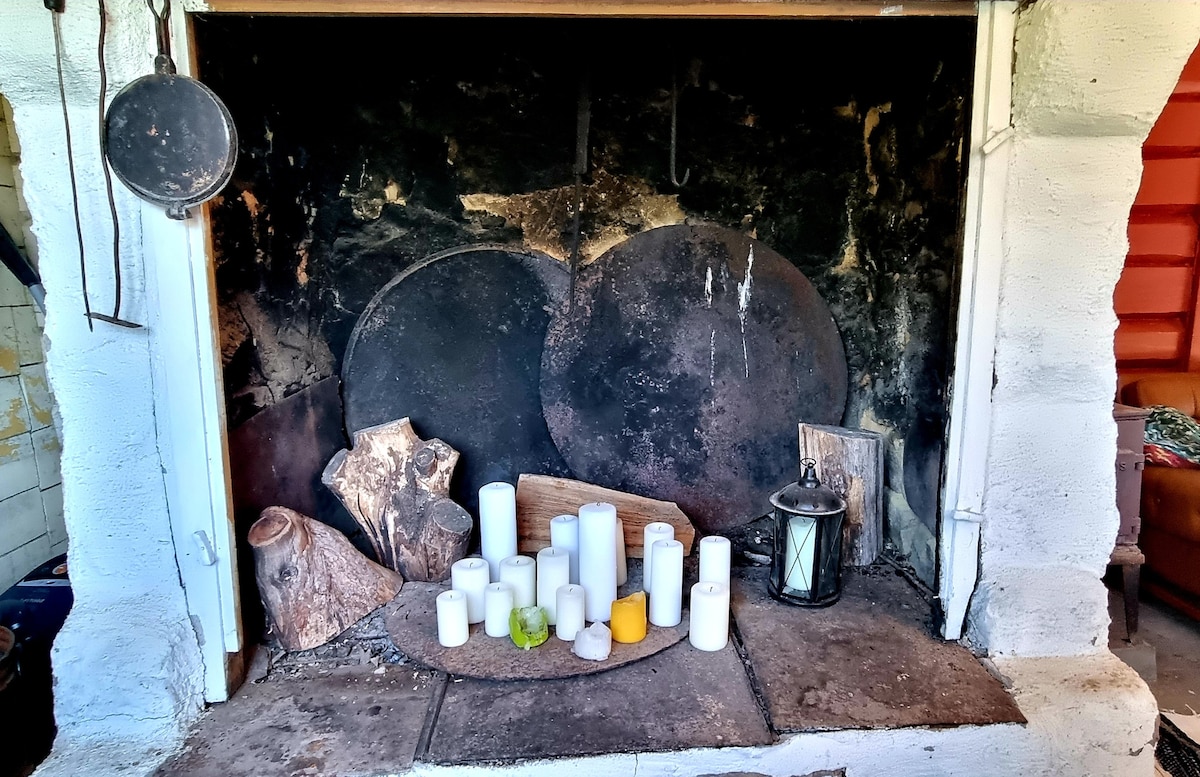 An old fireplace is showcased, featuring a mix of large round metal surfaces alongside various objects including wooden logs and candles. A lantern adds a touch of warmth to the rustic arrangement, while the soot-stained walls reflect its long history.