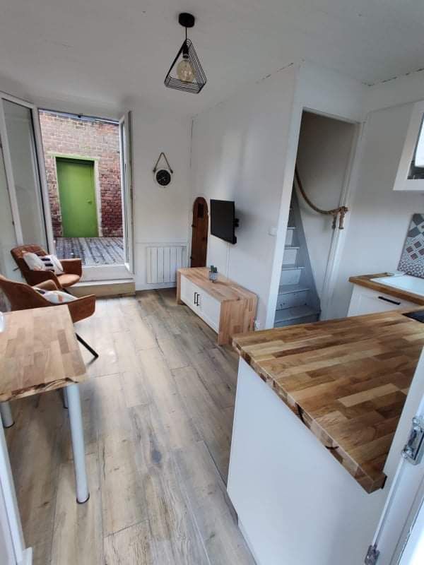 The living area features light wood flooring and a modern kitchenette with a wood countertop. A wall-mounted television is positioned across from a comfortable chair. Natural light enters through large windows, leading to a private outdoor terrace, visible through an open door.