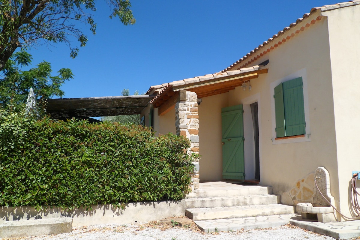 The exterior of a small independent house is displayed, featuring a stone entrance with steps leading up to the door. Green shutters frame the windows, and lush greenery surrounds the home. A shaded area is seen to the side, offering a serene outdoor space.