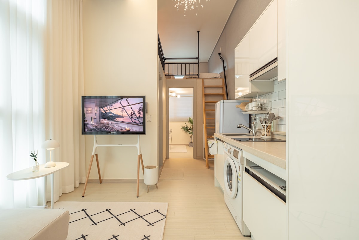 A bright living area is featured with a white couch and a small coffee table. A mounted television displays an image above a stylish kitchenette. Natural light is allowed in through large windows with sheer curtains, enhancing the spacious feel of the space.