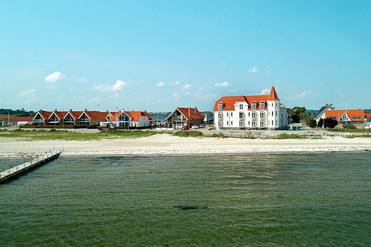 A gentle shoreline is framed by calm waters, with the white facade of Hejlsminde Badehotel visible against a clear blue sky. The hotel’s unique architecture features a red-tiled roof, surrounded by charming beach houses, contributing to the serene seaside ambiance.