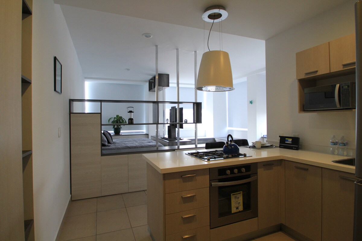 A modern kitchen space is visible, featuring light wood cabinetry and stainless steel appliances. A gas stove with four burners and a hood is present, along with a microwave and refrigerator. The open layout connects to a cozy sleeping area with a bed and subtle natural light.